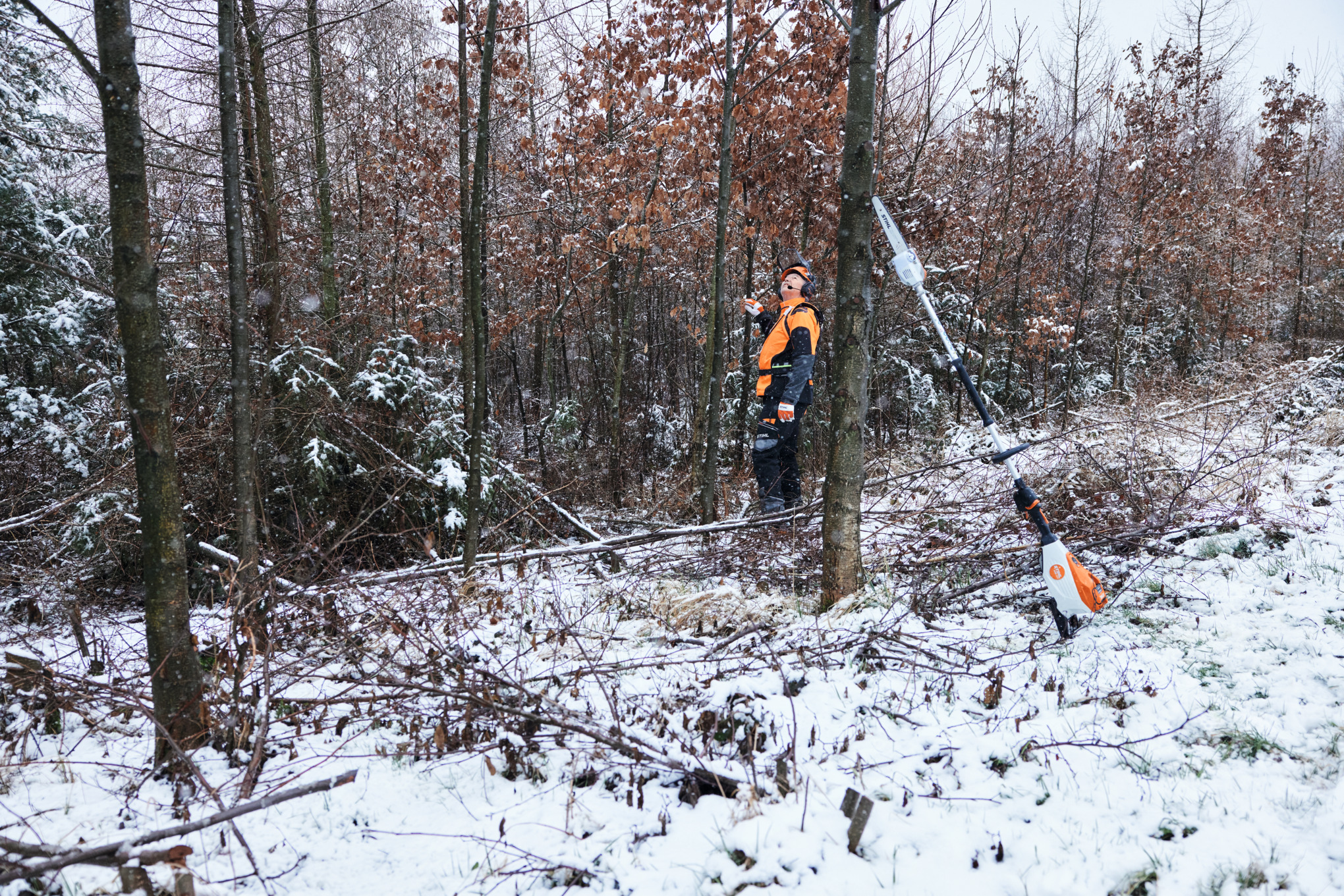 En mann i verneutstyr som arbeider i en snøkledd skog, mens en STIHL HTA 150 batteridrevet grensag står lent opp mot en trestamme i forgrunnen