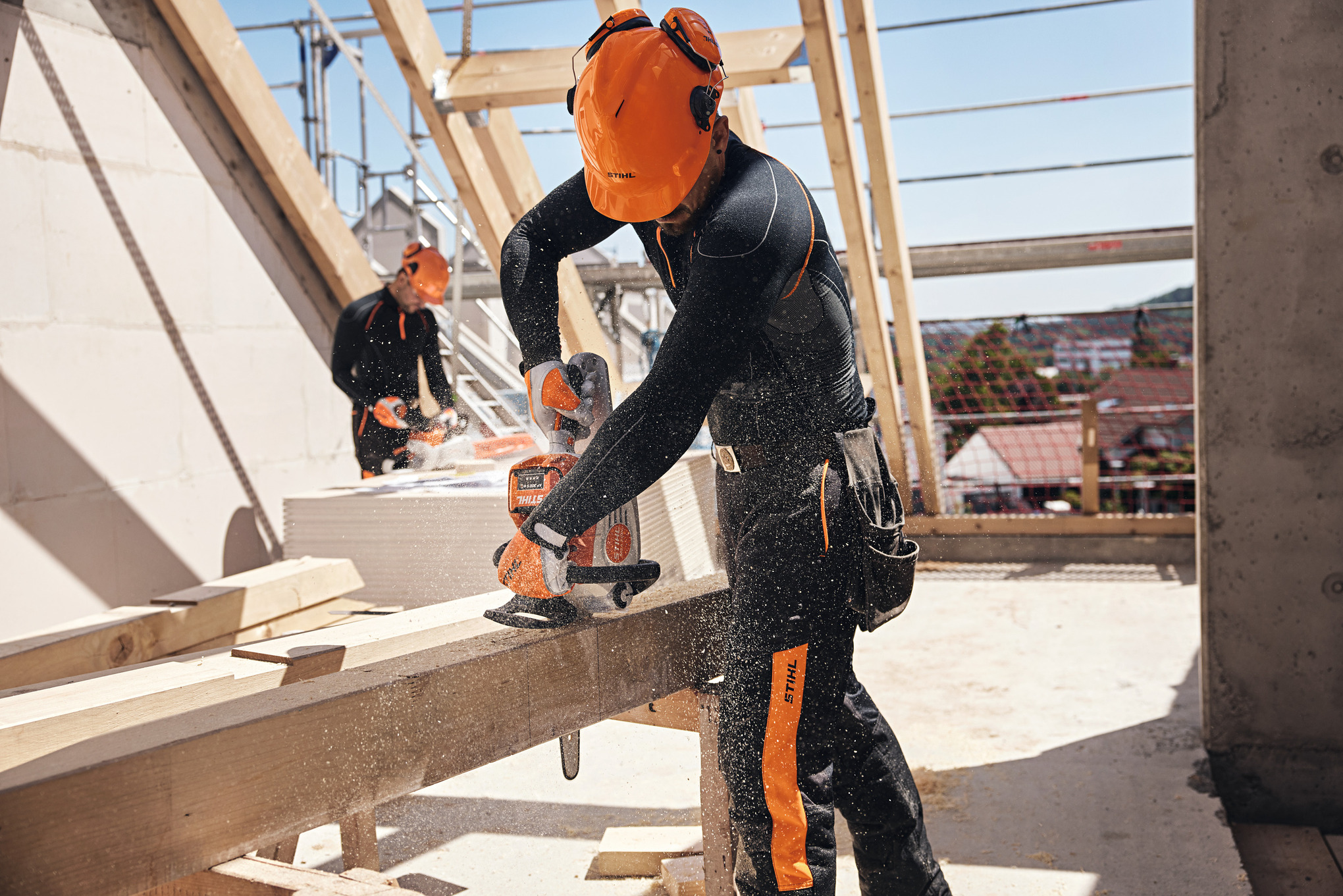En fagperson fra STIHL sager en trebjelke med en batteridrevet motorsag på et loft, mens en kollega bruker en batteridrevet motorsag i bakgrunnen