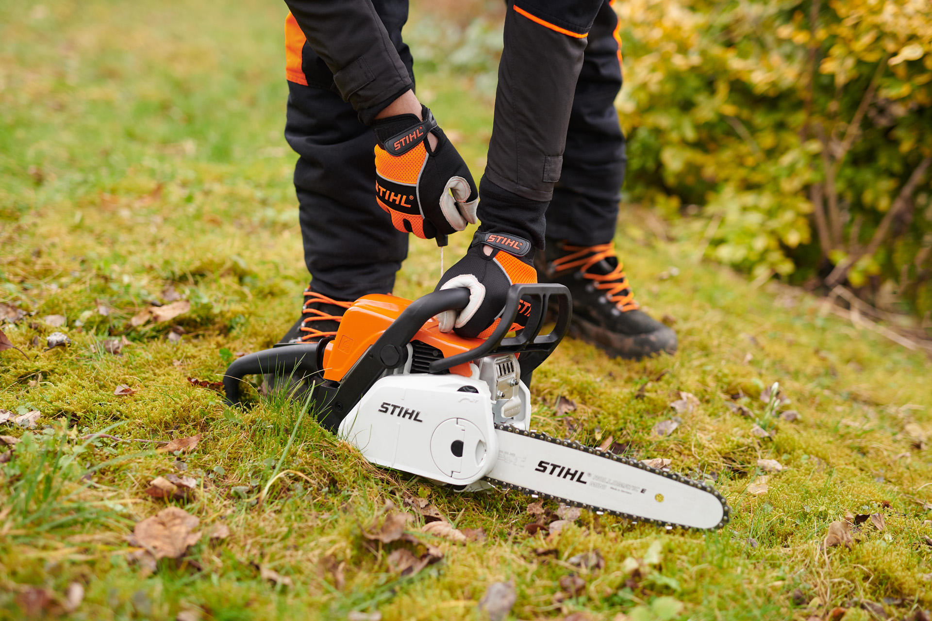 En STIHL MS 180 C-BE motorsag på bakken, mens en person holder i håndtaket med vernehansker.