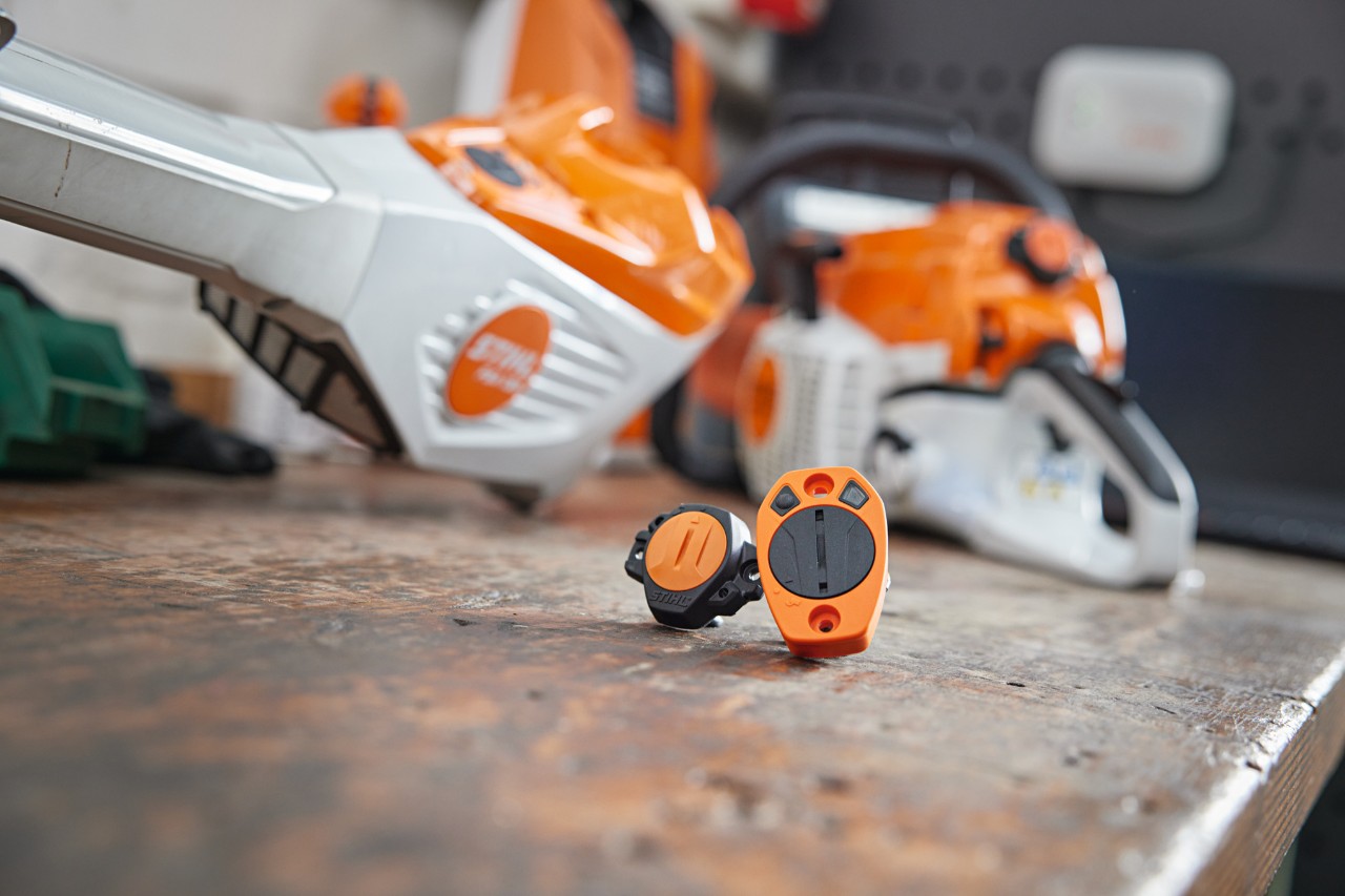 En STIHL Smart Connector og en STIHL Smart Connector 2 A på en arbeidsbenk. Forskjellige STIHL-redskaper ses uklart i bakgrunnen.