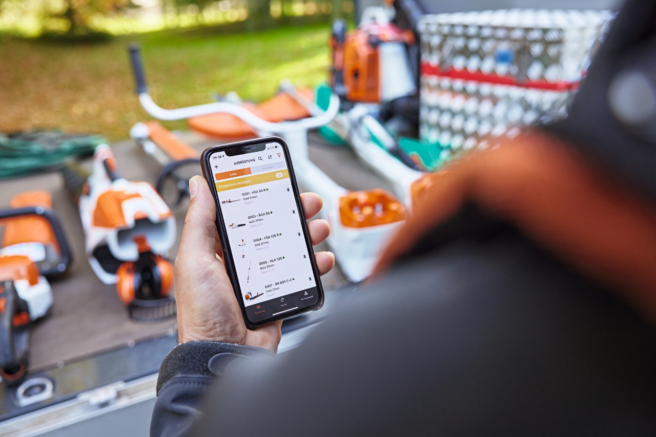 En person står foran lasteplanet til et kjøretøy som det står forskjellige STIHL-verktøy på. Personen holder en smarttelefon i hånden med STIHL Connected-appen åpen. 