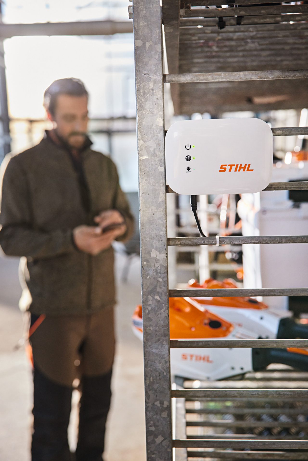 En STIHL Connected Box festet til en verkstedhylle. I bakgrunnen er det en anleggsgartner som bruker smarttelefonen sin. 