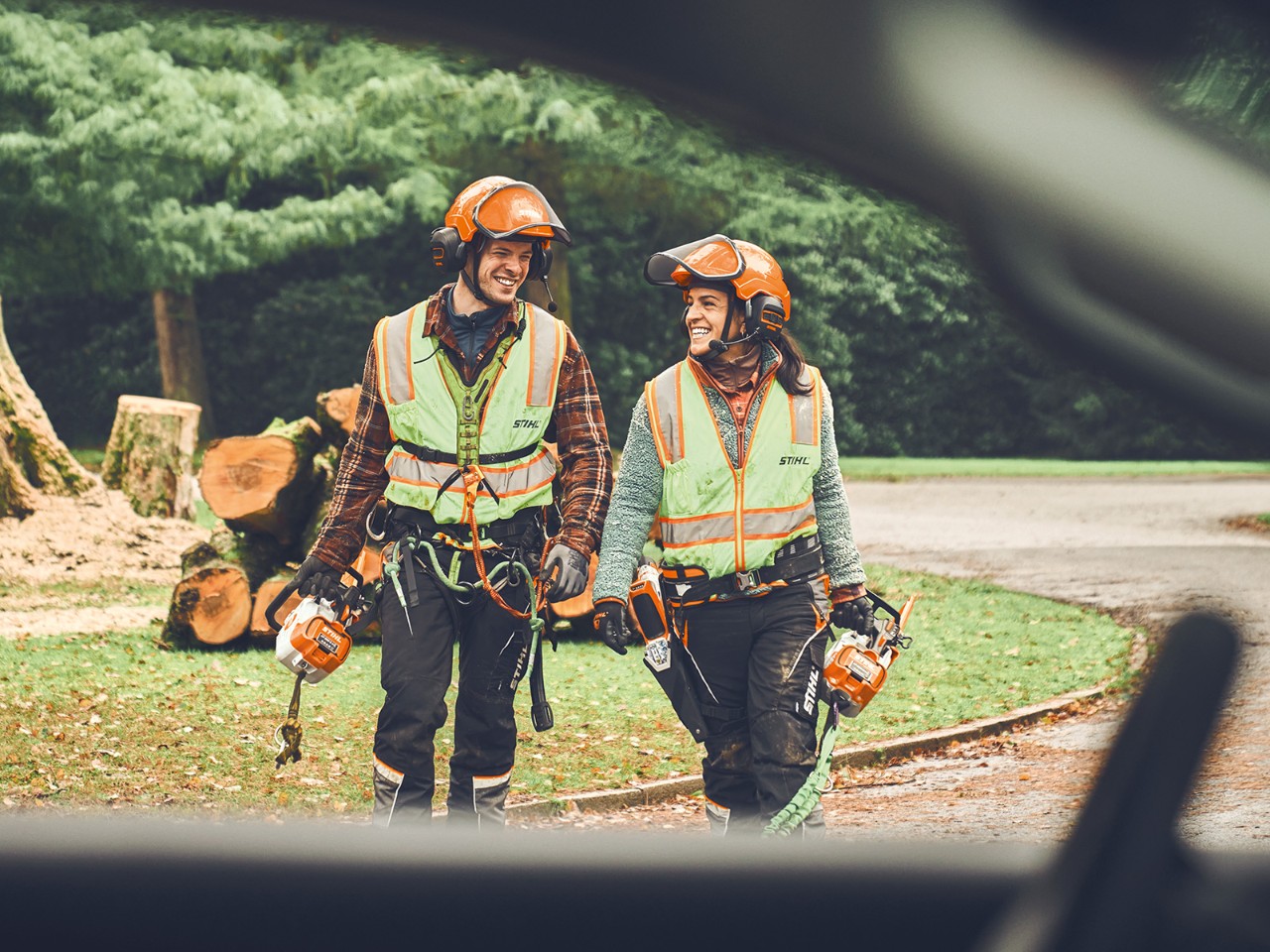 En mann og en kvinne med STIHL batteridrevne motorsager og en STIHL trådløs tretrimmer går mot en bil, smilende