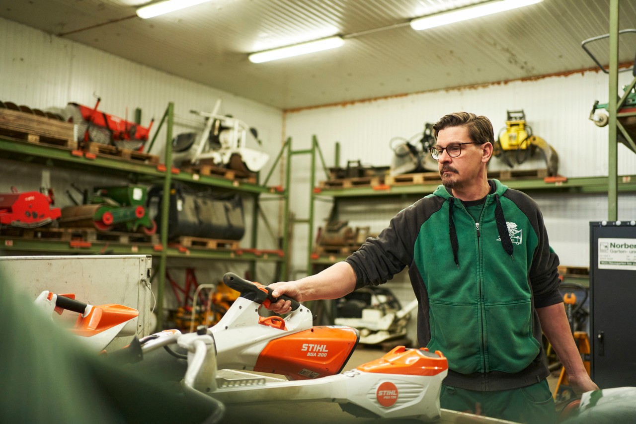 En mann i et verksted står ved en benk med STIHL batteridrevne verktøy og griper tak i en STIHL BGA 200 batteridrevet løvblåser