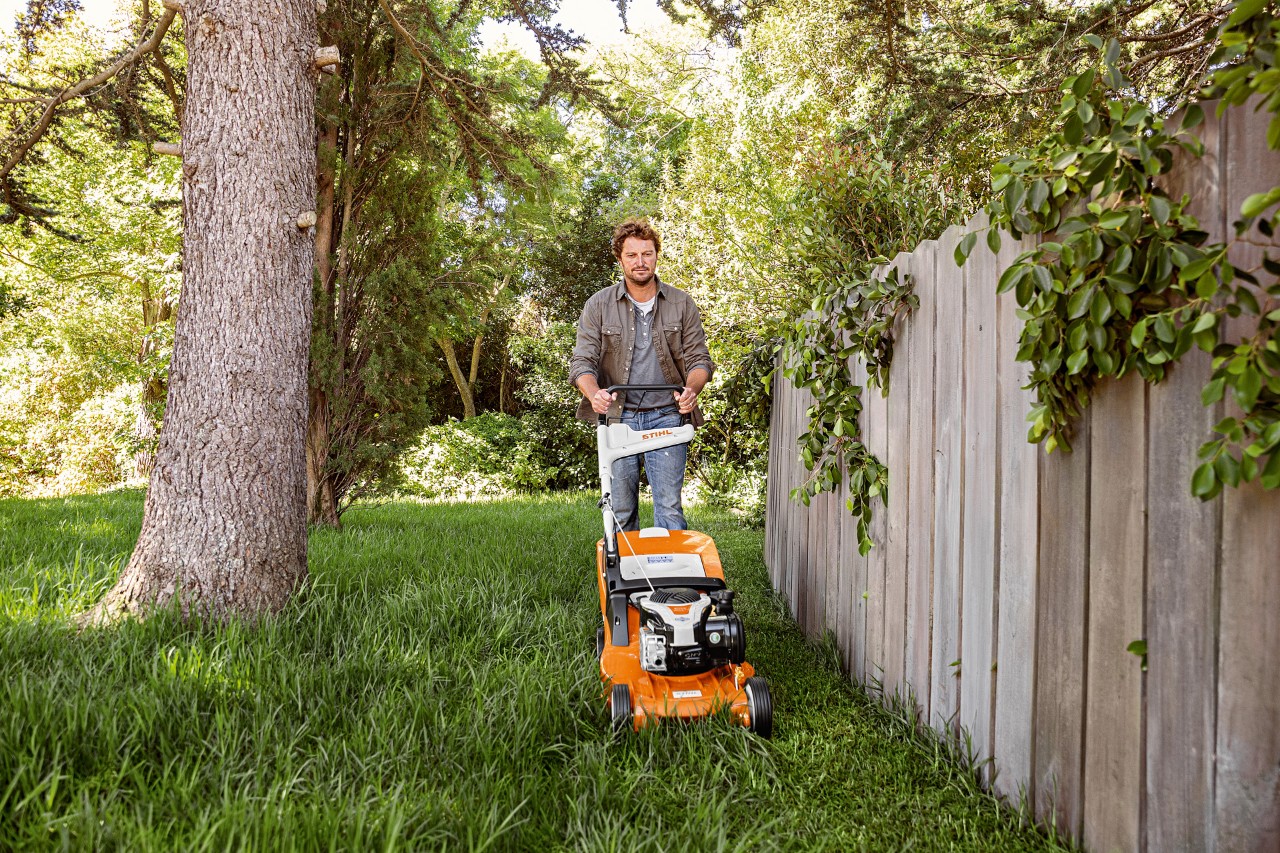 En mann klipper plena med en bensin-gressklipper RM 448 TC fra STIHL ved siden av et tregjerde