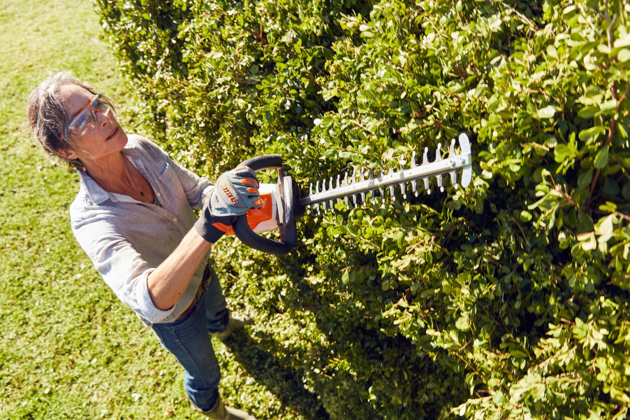 En kvinne med vernebriller trimmer en høy hekk med en batteri-hekksaks HSA 56 fra STIHL