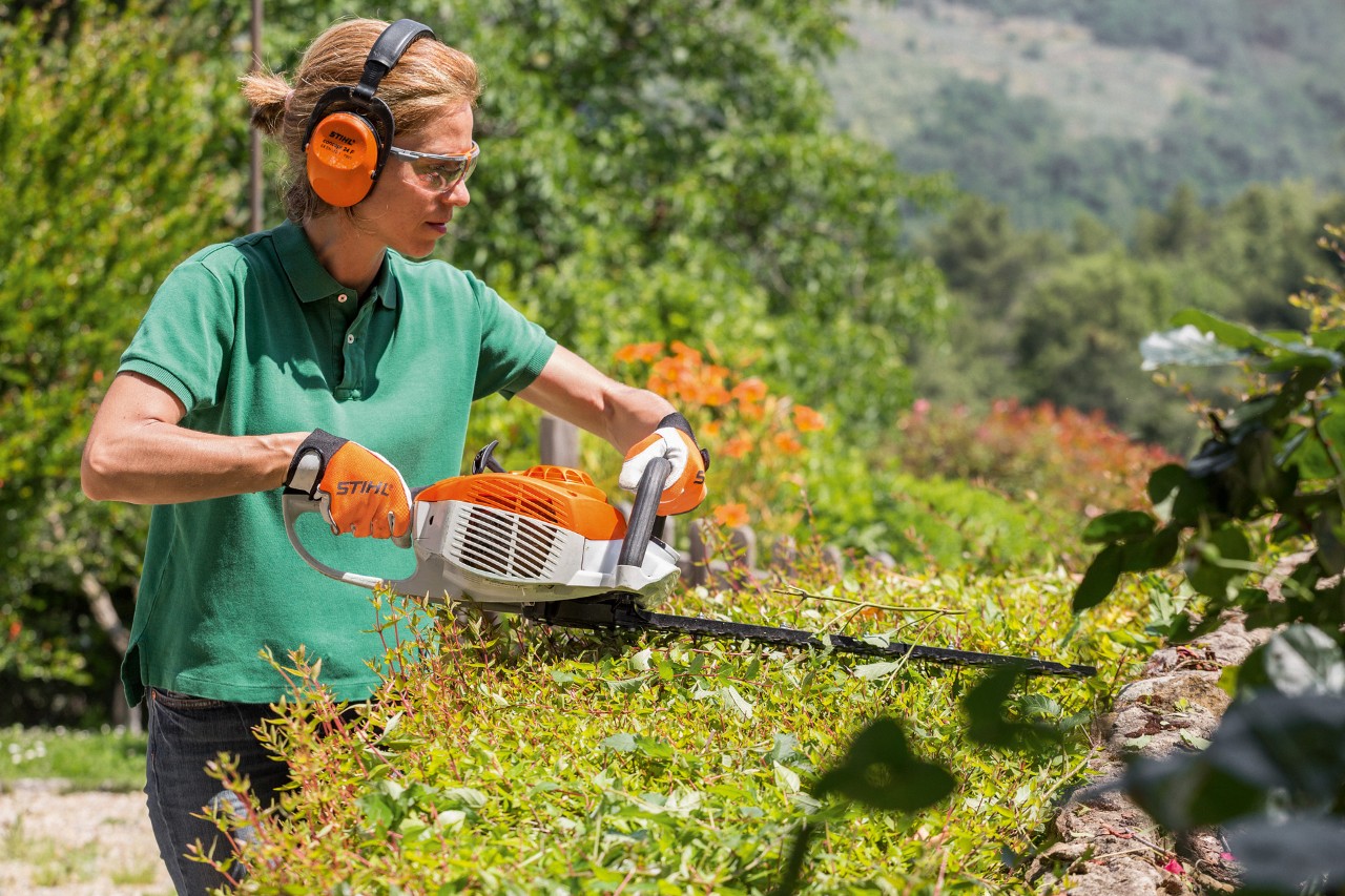 En kvinne med hørselvern trimmer en hekk med bensin-hekksaksa HS 46 fra STIHL