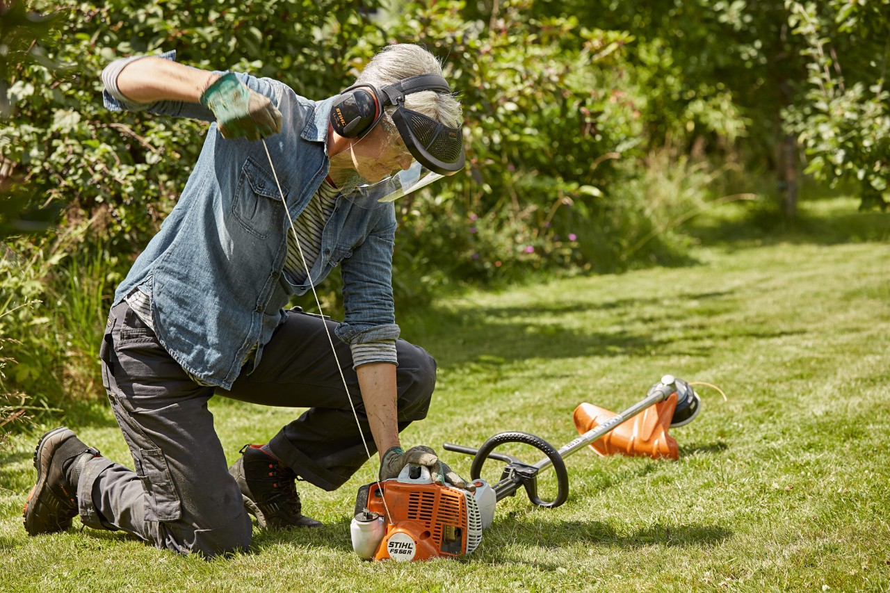 Kvinne med hørselsvern og ansiktsvern som starter en STIHL FS 56 R bensindrevet ryddesag i nærheten av gress
