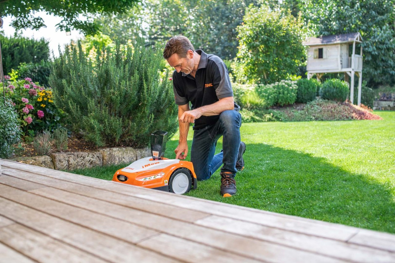 En mann kneler ved siden av en STIHL iMOW® robotgressklipper i en hage med busker og en terrasse