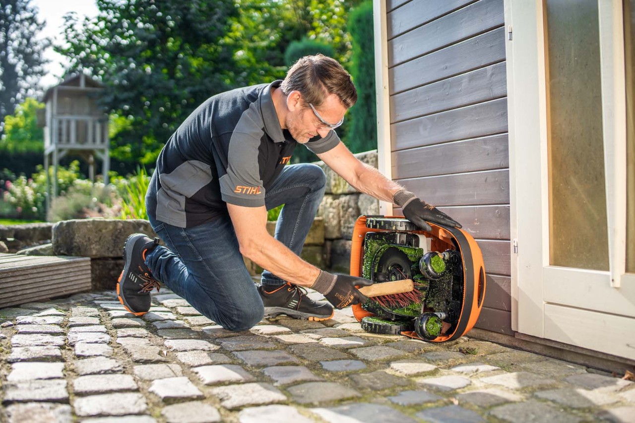 En mann rengjør undersiden av en STIHL iMOW® 422 robotgressklipper med en kost 