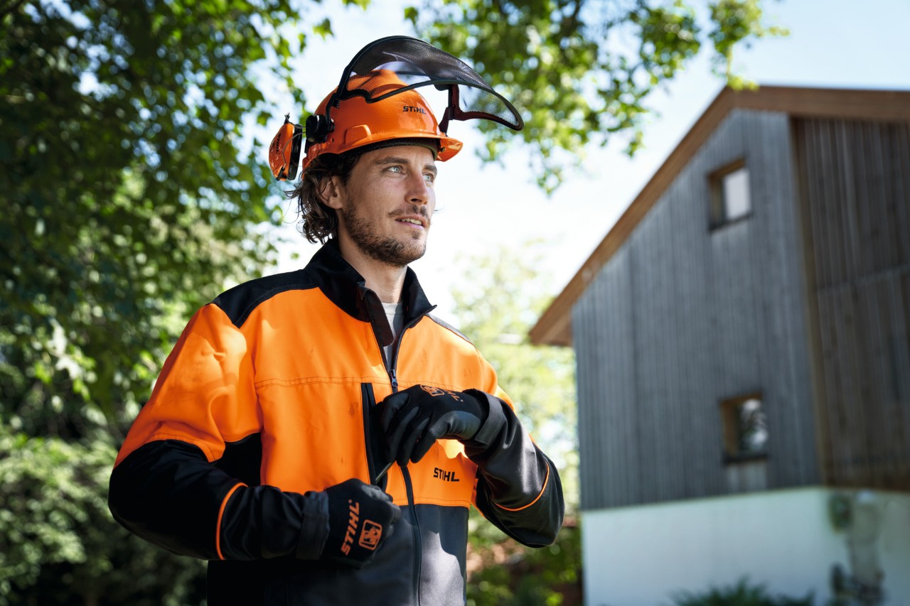 En mann med oransje og svart verneutstyr fra STIHL, blant annet hjelm og hansker, stirrer ut av bildet med et beroligende ansiktsuttrykk, og med hus og trær i bakgrunnen 