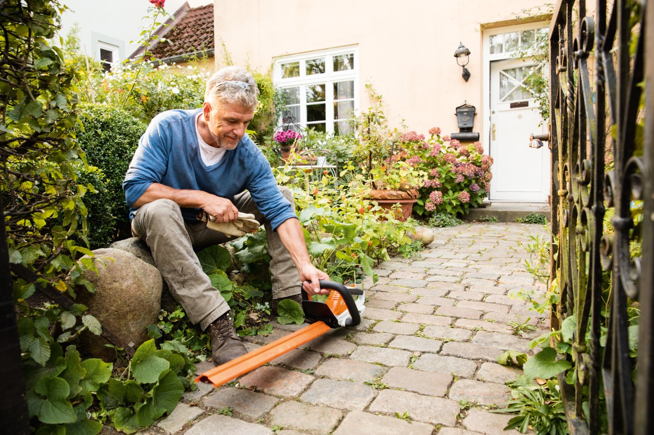 En mann sitter på en stor stein med en HSA 45 elektrisk hekksaks fra STIHL AI-serien foran et hus