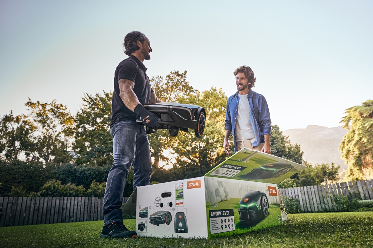 En STIHL-forhandler står i hagen til en kunde med en iMOW® EVO i hånden og iMOW®-boksen foran seg, klar til å hjelpe til med installasjonen