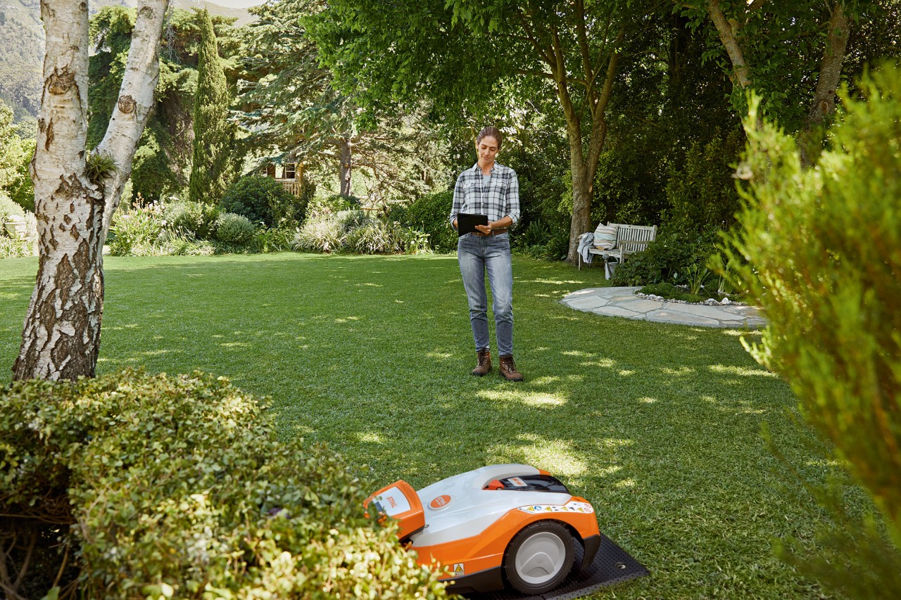 En kvinne bruker et nettbrett i hagen og betjener en STIHL iMOW® 522 robotgressklipper